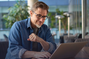 A man wearing glasses smiles at his laptop