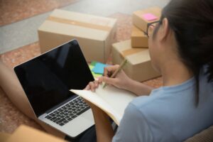 A young woman makes notes in a book in front of a blank laptop and some boxes ready to ship.